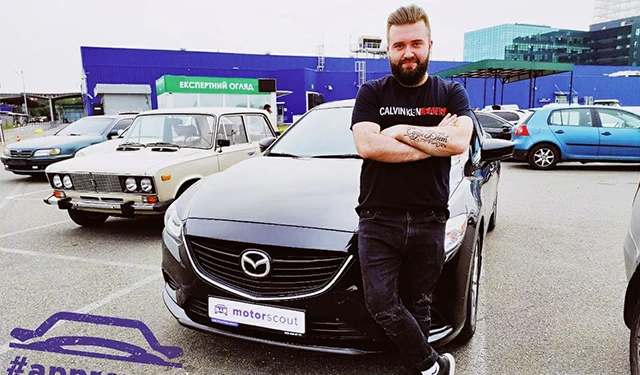 Чоловік поруч із чорним автомобілем на парковці з іншими машинами.